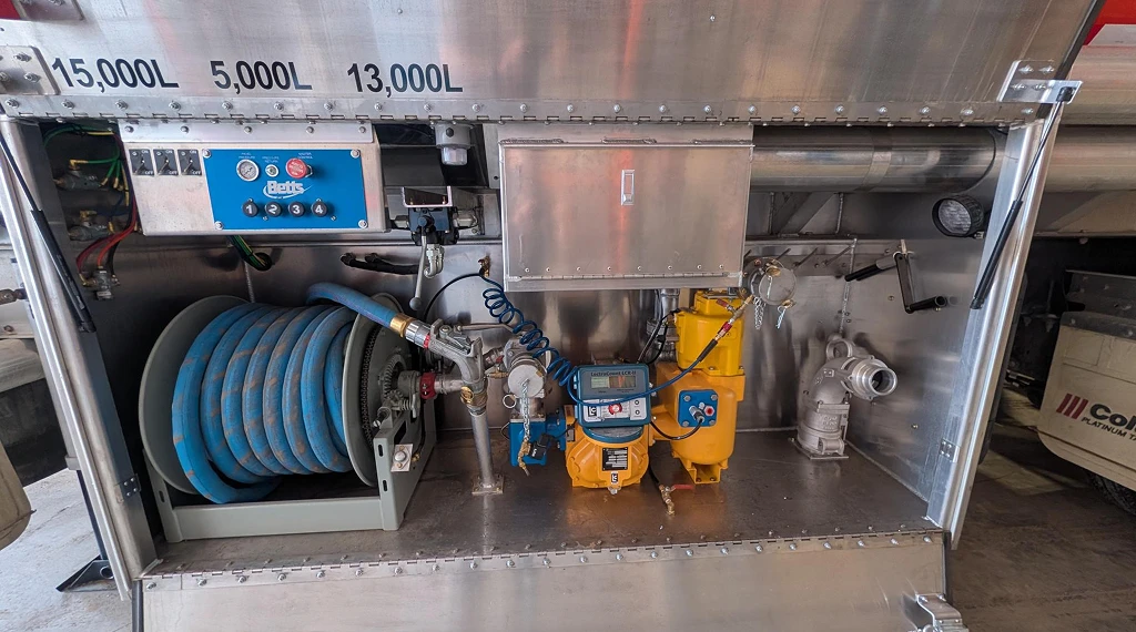 Close-up of a fuel delivery truck pump system with hose reel, controls, and fuel compartments.