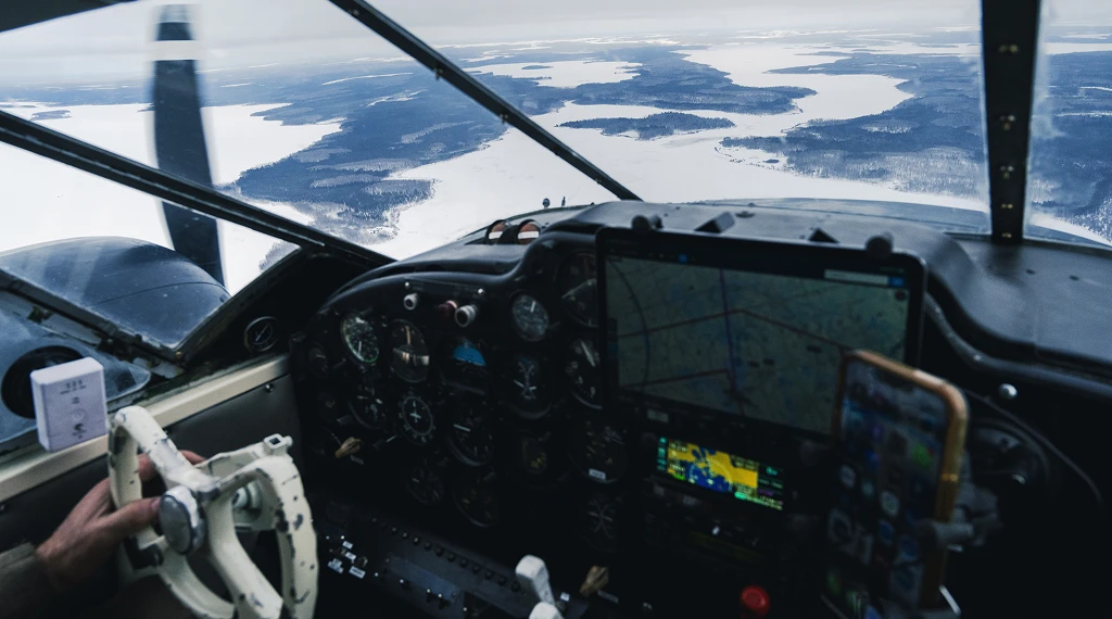 View from inside a small aircraft cockpit flying over a remote snowy landscape.